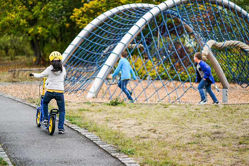Ein Mädchen fährt im Garten Roller, zwei Jungen spielen Fangen.