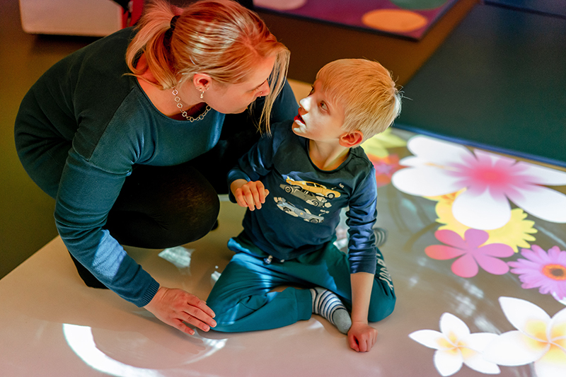 Ein Junge und eine Frau sitzen auf einem weißen Boden, auf den Blumenbilder projeziert werden.