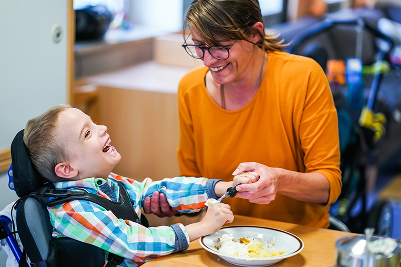 Eine pädagogische Fachkraft reicht einem kleinen Jungen im Rollstuhl das Essen