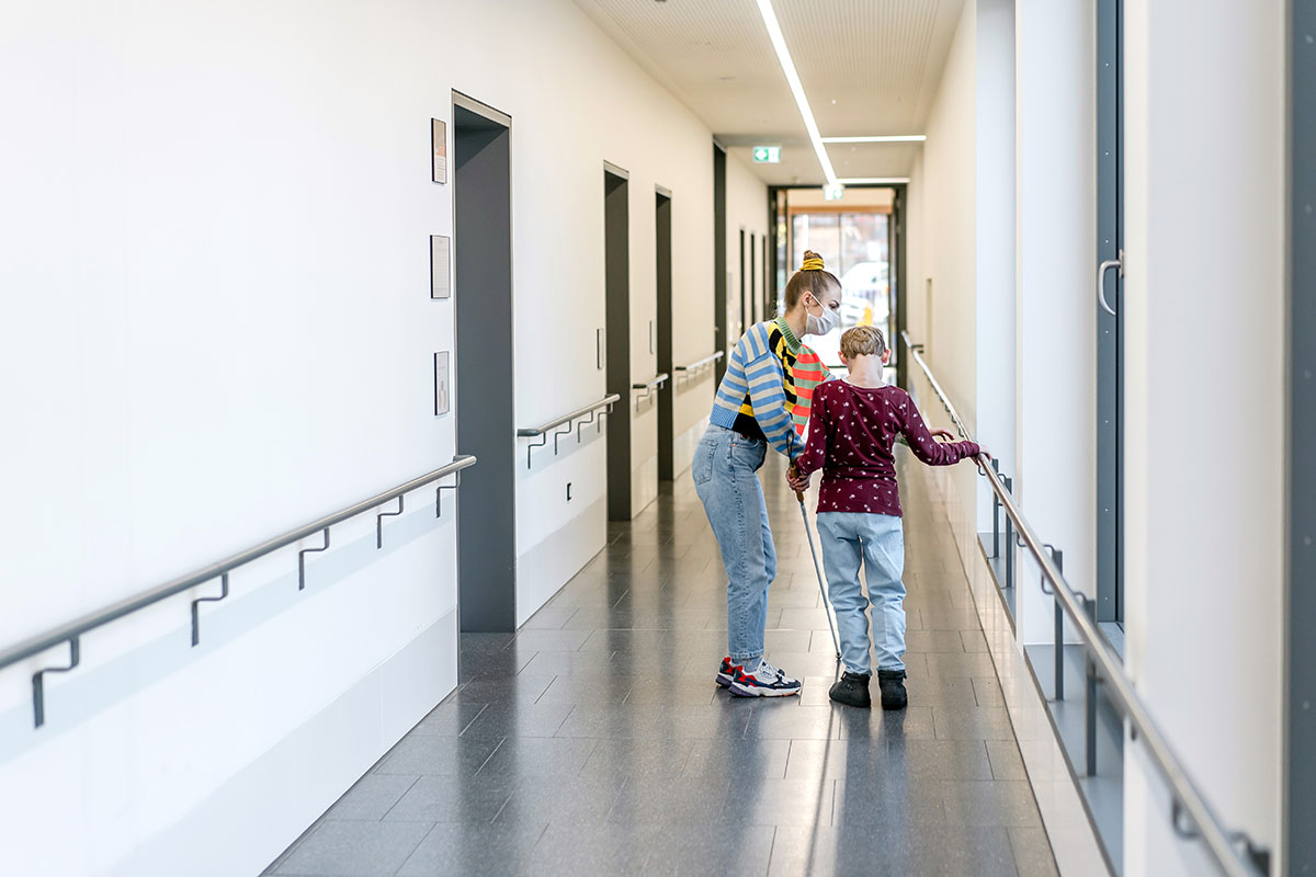 Eine Frau und ein Kind stehen im Schulflur. Das Kind trägt einen Blindenstock und hält sich an einem Handlauf fest, welcher an den Wänden angebracht ist.
