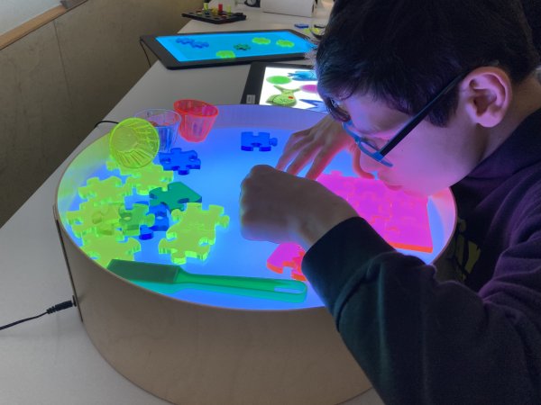 Auf dem Bild sieht man einen jungen Mann, der sich über einem Leuchtkasten beugt. Darauf liegen Puzzleteile, mit denen er spielt.