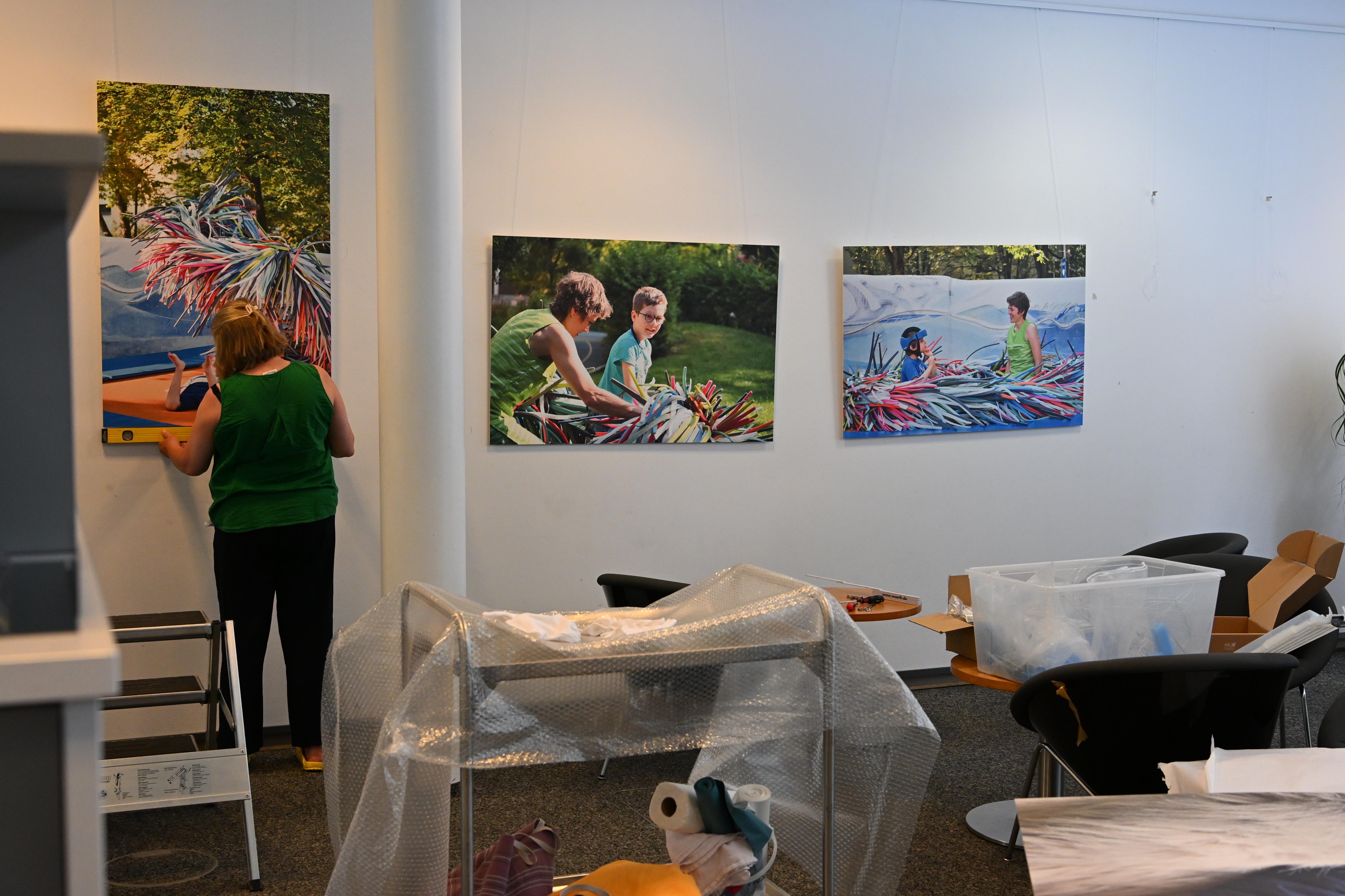 Auf dem Bild sieht man eine Frau von hinten, wie sie an einer Wand, an der bereits Bilder hängen, weitere Bilder aufhängt.
