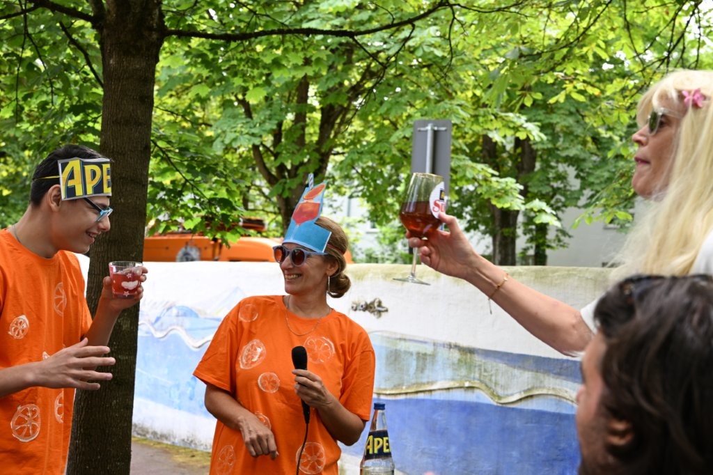Auf dem Bild siehtman zwei Personen mit orangem T-Shirt und einer Aperol-Mütze, sowie eine blonde Frau. Alle drei stoßen mit alkoholfreiem Aperol miteinander an.