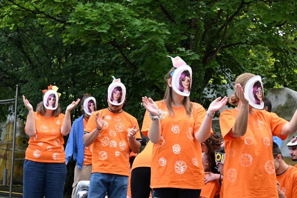 Auf dem Bild sieht man mehrere Personen mit orangen T-Shirts und einer Pappmaske mit einem Gesicht drauf.