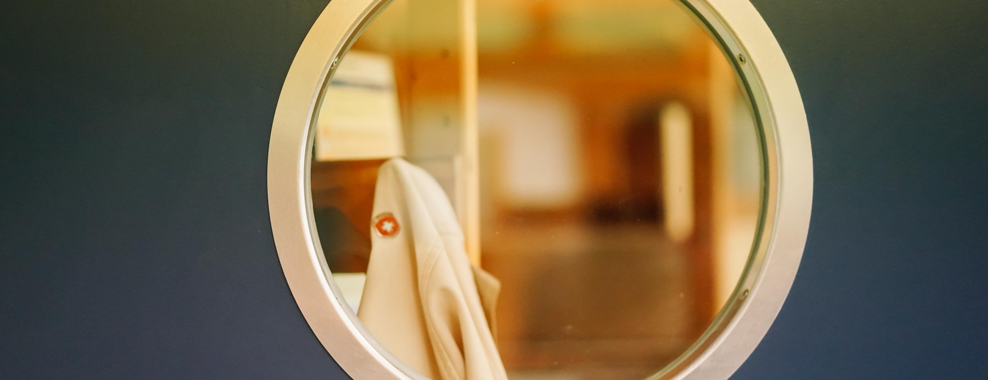 Ein rundes Guckloch aus Glas in einer grauen Wand. Dahinter sind unscharf braune Gegenstände zu erkennen.