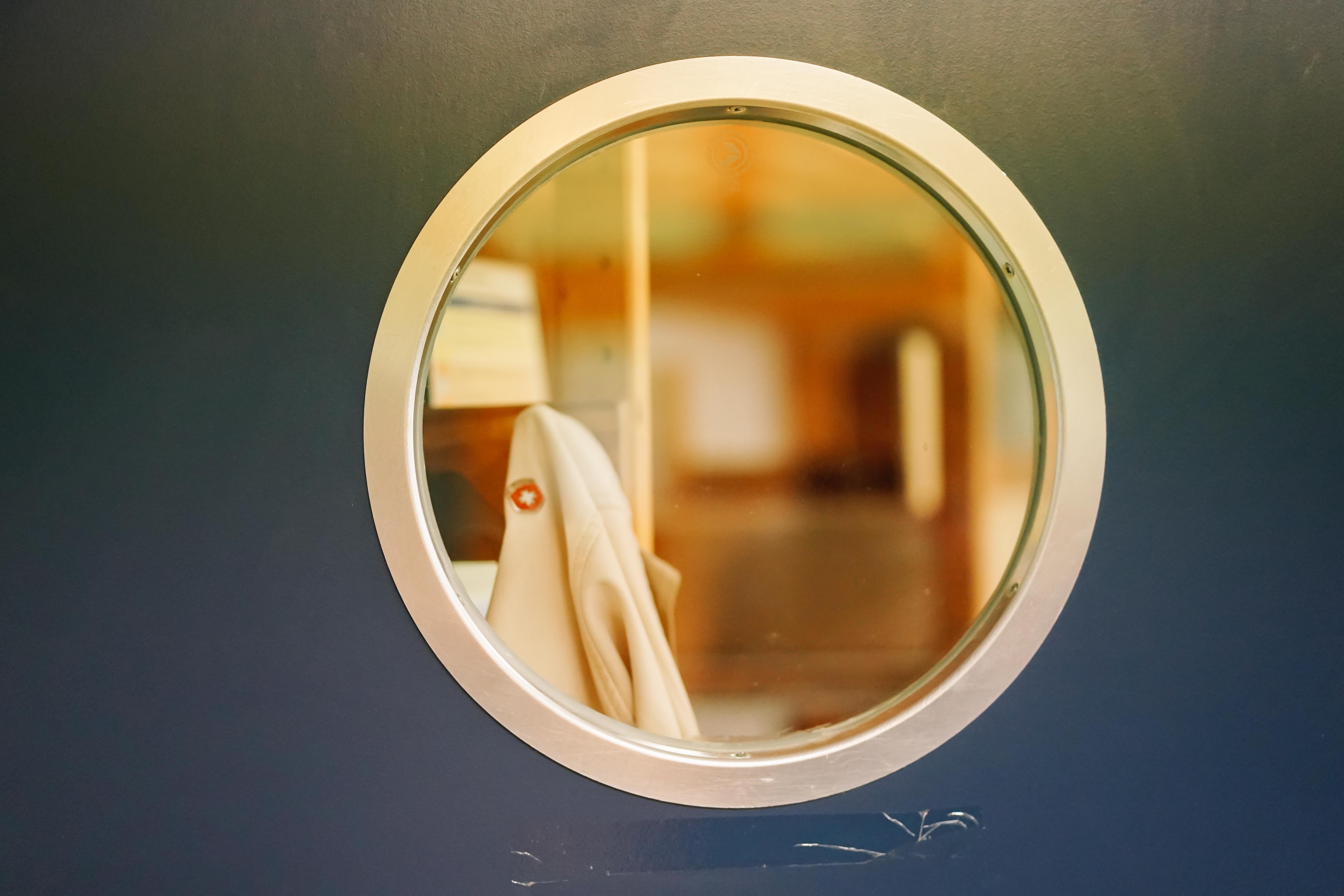 Ein rundes Guckloch aus Glas in einer grauen Wand. Dahinter sind unscharf braune Gegenstände zu erkennen.