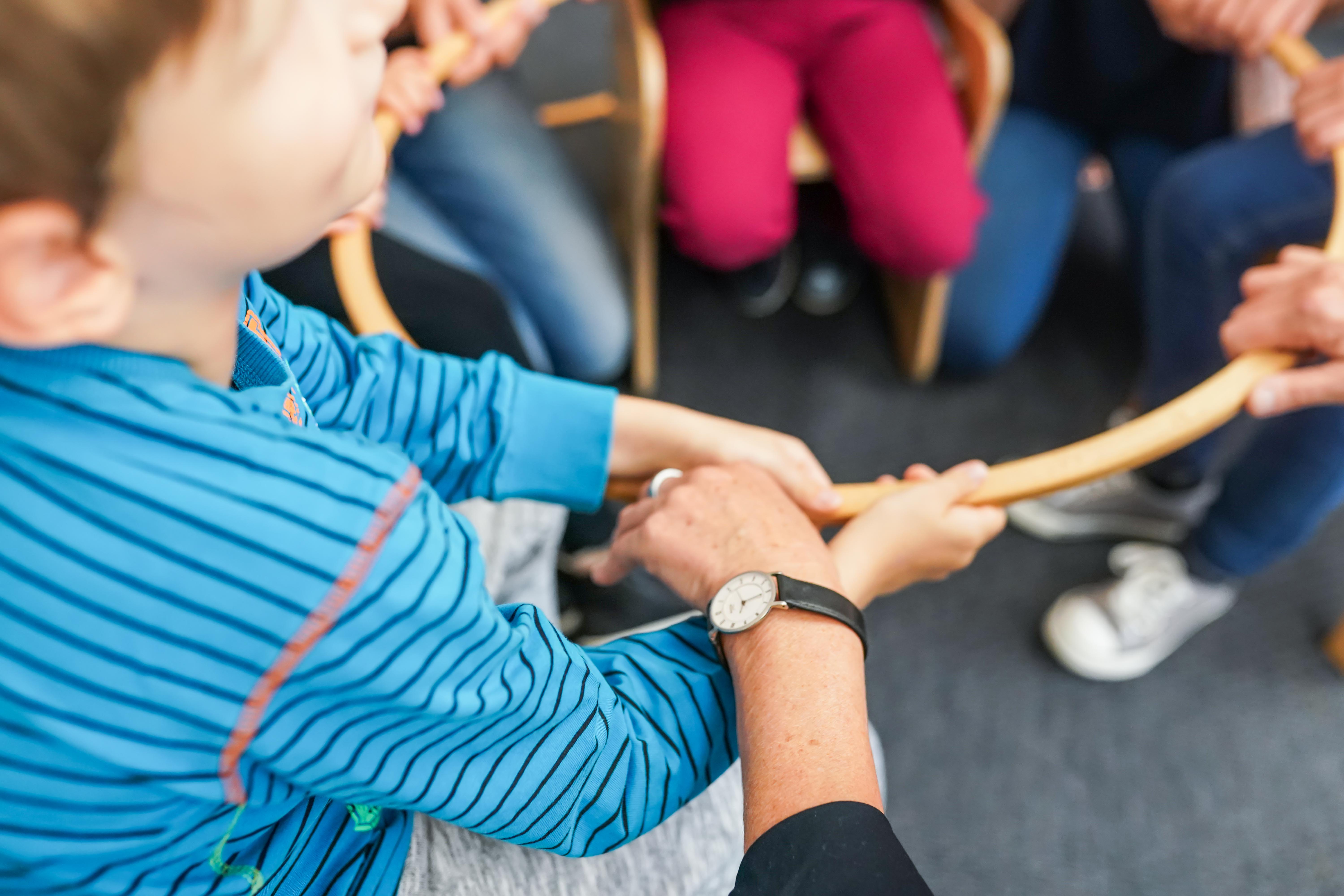 Mehrere Personen, darunter ein Kind im Vordergrund, halten gemeinsam einem Hula Hoop  aus Holz fest. Das Kind trägt ein blaues Shirt mit dunklen Streifen. Nur die Hände und Arme der anderen Personen sind sichtbar. Sie sitzen auf Stühlen. Die Szene wirkt wie ein Gruppenspiel oder eine gemeinsame Aktivität, bei der Koordination und Zusammenarbeit gefragt sind. Eine erwachsene Hand mit Armbanduhr unterstützt das Kind beim Halten des Hula Hoops.