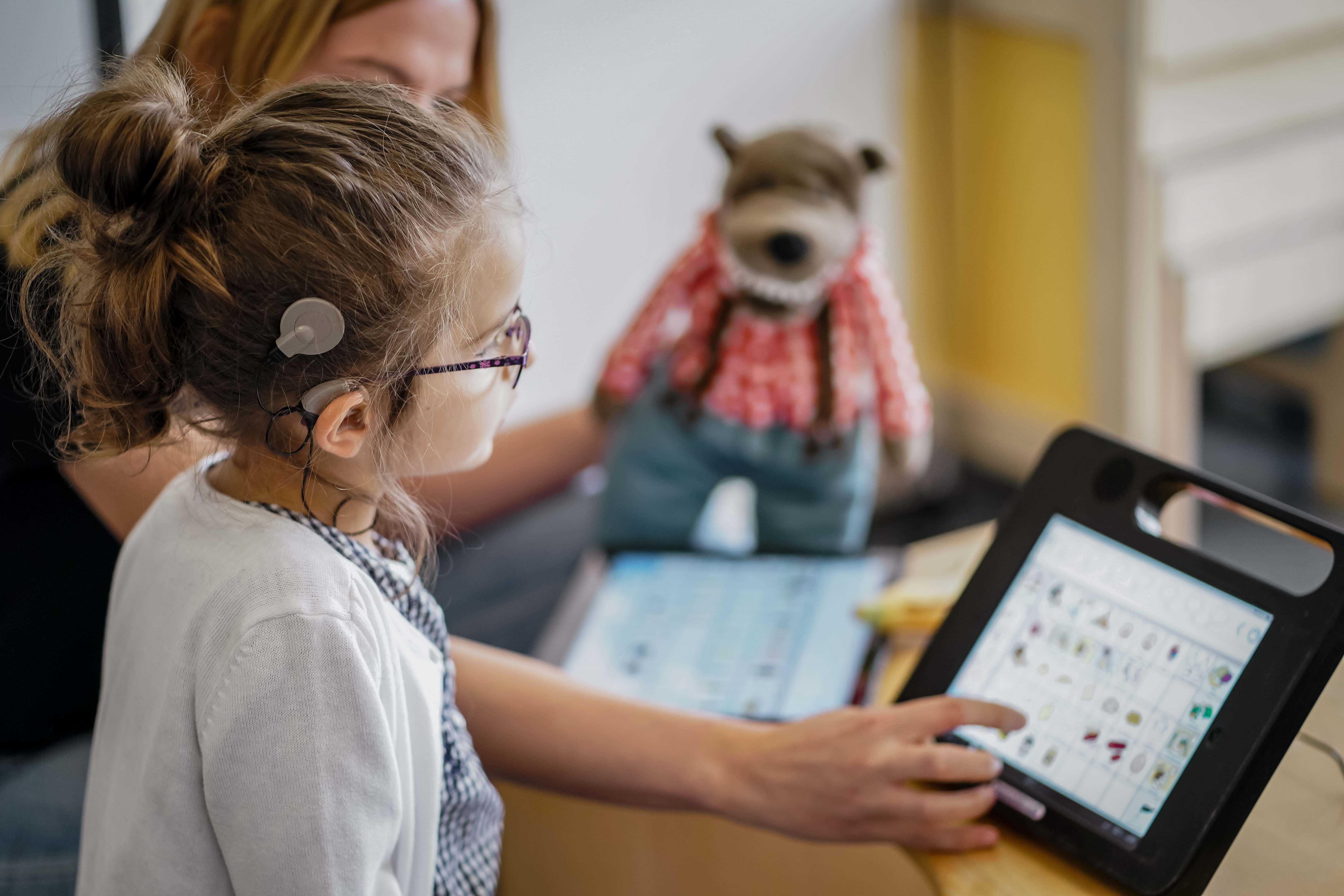 Ein Kind mit einem Cochlea-Implantat sitzt an einem Tisch und schaut auf ein Tablet, auf dem Symbole und Bilder zu sehen sind. Vermutlich handelt es sich um eine unterstützende Kommunikations-App. Im Hintergrund ist eine erwachsene Person zu erkennen, die auf das Tablet zeigt. Auf dem Tisch, auf dem das Tablet steht, sitzt außerdem ein Stofftier. Die Szene wirkt wie ein inklusives Lern- oder Therapieumfeld, in dem das Kind mithilfe digitaler Hilfsmittel kommuniziert.