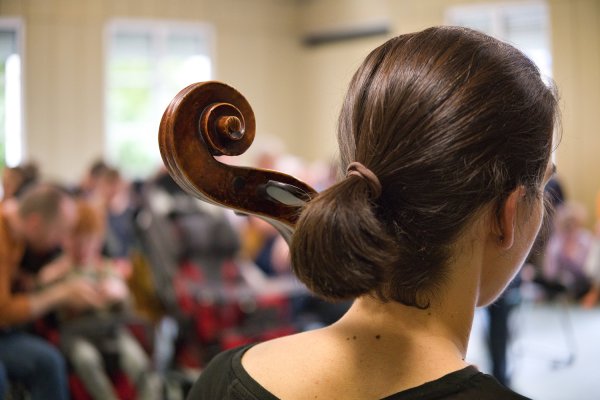 Das BIld zeigt eine Cello-Spielerin von hinten. Im Hintergrund sieht man Publikum.