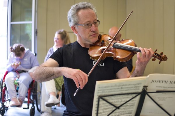 Auf dem Bild sieht man einen Geiger des BRSO spielen; im Hintergrund sieht man zwei Personen, die zuhören.