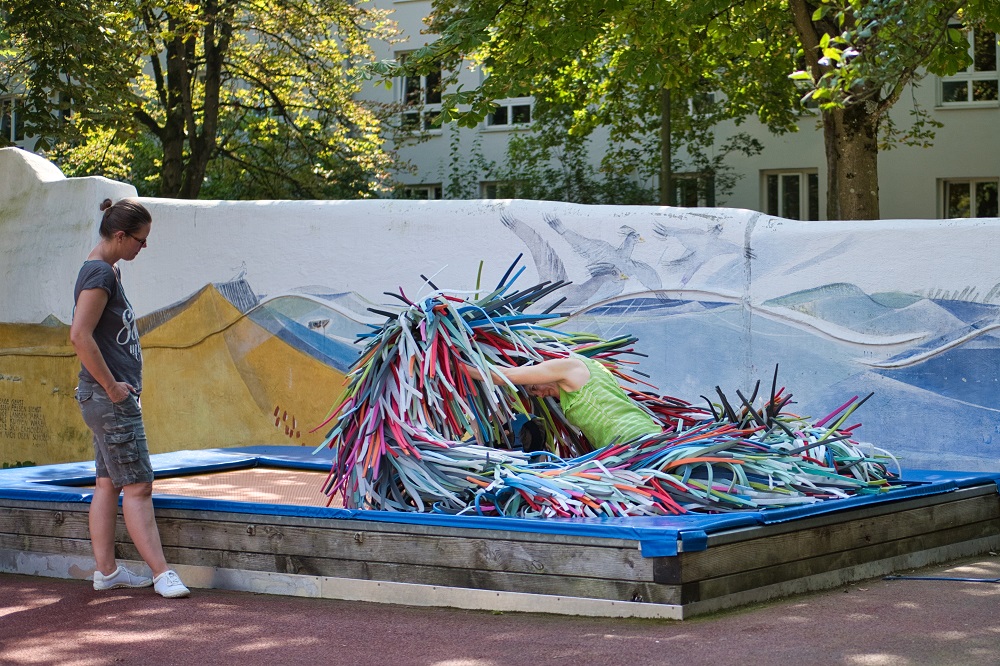 Das Bild zeigt eine Frau neben einem Trampolin. Auf dem Trampolin befindet sich die Kunstfigur ZWINK bestehen aus bunten Plastikschlangen.