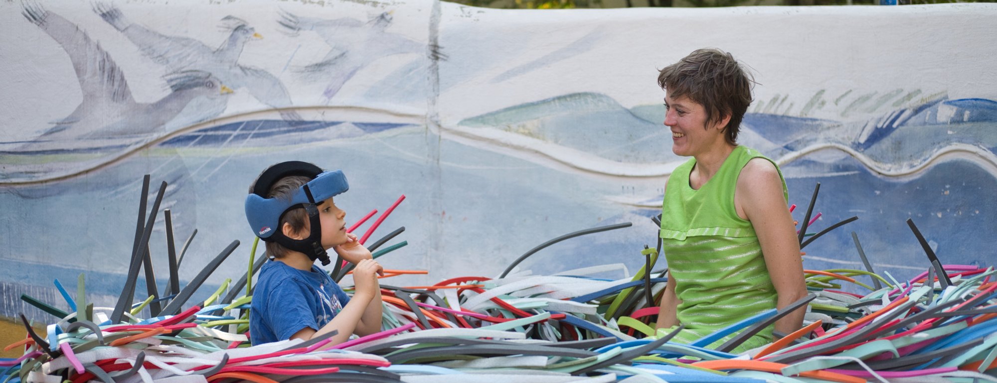 Das Bild zeigt einen Jungen und eine junge Frau. Die beiden sitzen auf einem Trampolin, drum herum das Kostüm ZWINK aus ganz vielen bunten Plastikstangen.