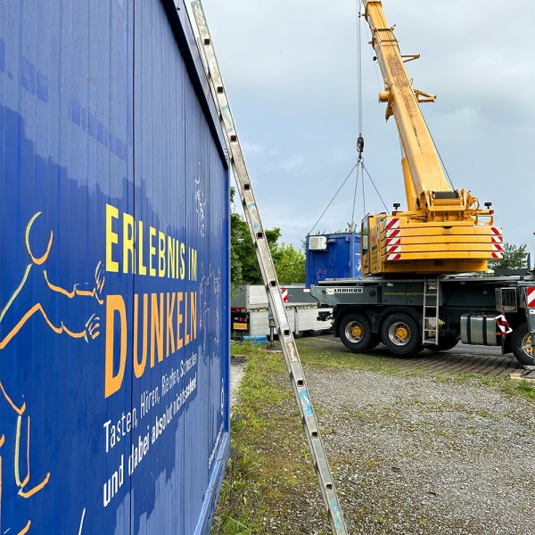 Im Vordergrund die blaue Wand eines Schiffscontainers mit der Aufschrift Erlebnis im Dunkeln. Im Hintergrund ein großer gelber Kran, der einen zweiten Container auf einen Hänger lädt.
