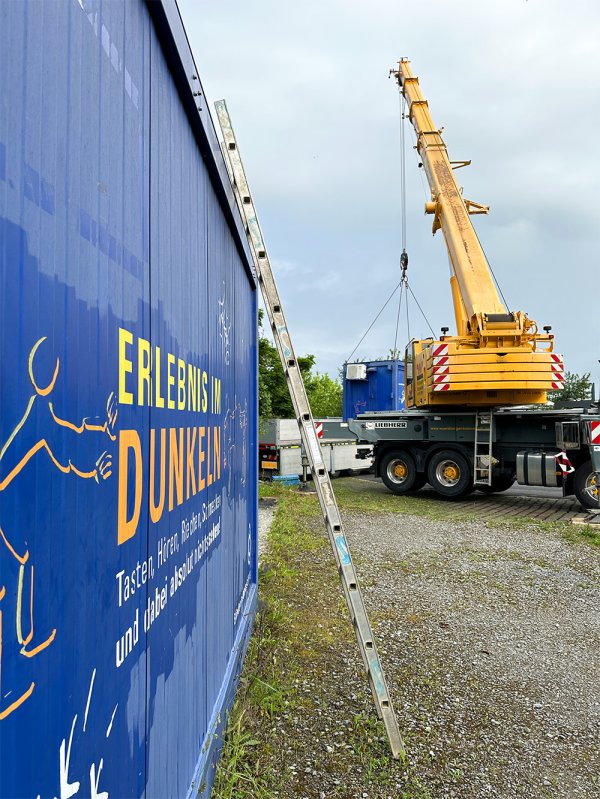 Im Vordergrund die blaue Wand eines Schiffscontainers mit der Aufschrift Erlebnis im Dunkeln. Im Hintergrund ein großer gelber Kran, der einen zweiten Container auf einen Hänger lädt.