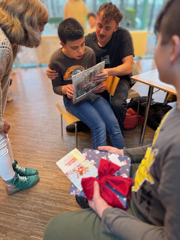 Mehrere Personen sitzen und stehen in einem Raum mit großen Fenstern. Eine Person hält ein verpacktes Geschenk. Eine andere Person daneben hält ein Geschenk mit rotem Band und eine Grußkarte in den Händen.