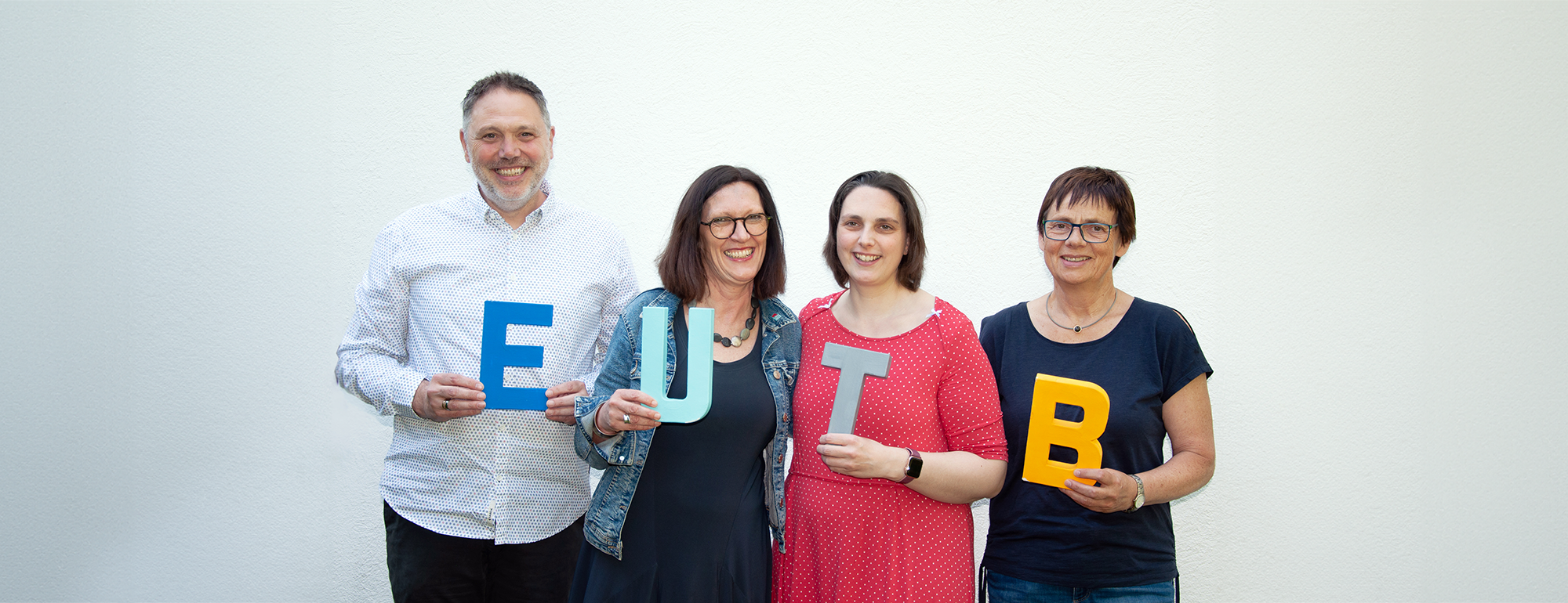 Ein Mann und drei Frauen stehen vor einer hellen Wand. Jeder von ihnen hält einen Buchstaben vor sich. Die Buchstaben sind E, U, T, B.