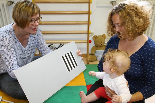 Links im Bild sitzt eine Orthoptistin mit einer Tafel zur Sehüberprüfung. Ihr gegenüber sitzt eine Mutter mit einem Kleinkind auf dem Schoß.