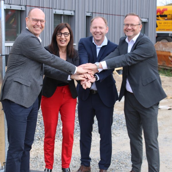 Gruppenbild: Drei Männer und eine Frau stehen vor einem grauen Gebäude und lachen in die Kamera. Sie haben ihre Hände symbolisch übereinander gelegt.