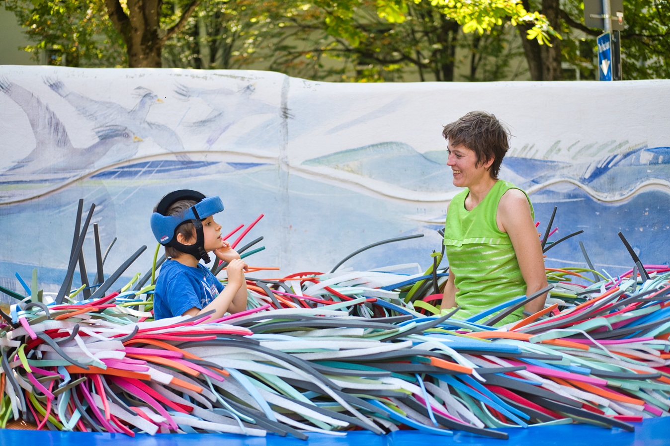 Das Bild zeigt einen Jungen und eine junge Frau, die auf einem Trampolin vor einer bunten Mauer sitzen.