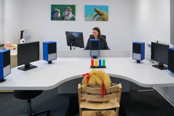 Eine Frau sitzt an einem Schreibtisch mit mehreren Monitoren und Lautsprechern. Im Vordergrund steht ein Holzstuhl mit einer Puppe davor, auf dem Tisch liegen farbige Zylinder.