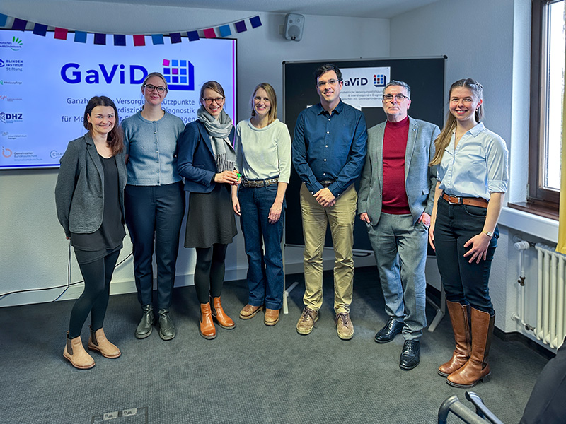 Sieben Personen stehen in einem Raum vor Bildschirmen mit der Aufschrift „GaViD“ und posieren für ein Gruppenfoto.