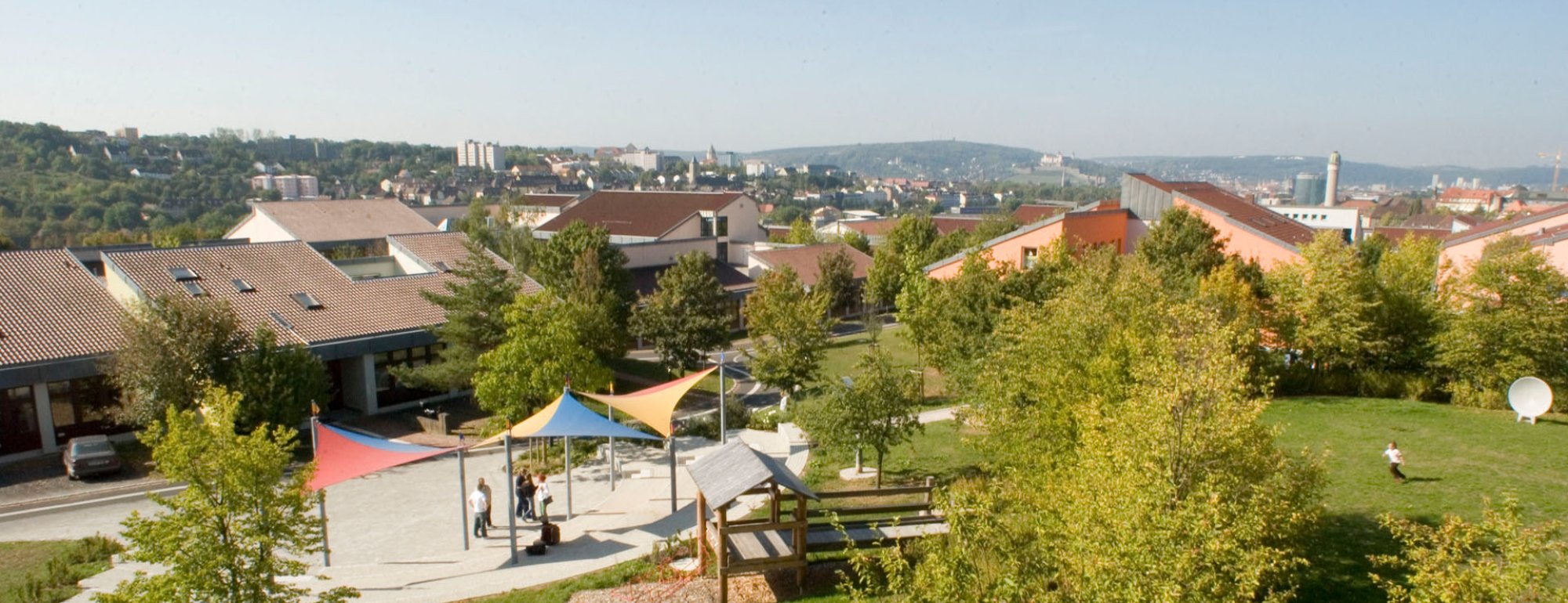 Neben einer grünen Wiese mit Spielgeräten und Bäumen ist ein gepflasteter Platz mit bunten Sonnensegeln. Im Hintgrund sind Häuser zu sehen, weiter hinten die Ausläufer einer Stadt und ganz kleine eine Burg.