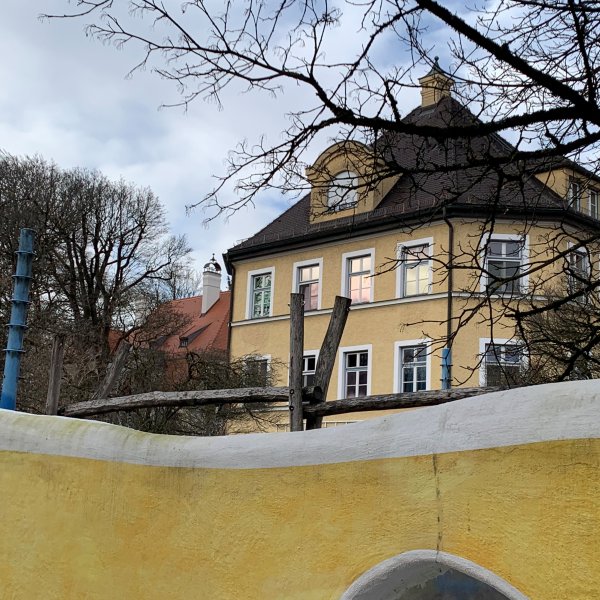 Das Bild zeigt eine gelbe Mauer und dahinter einen Teil des Gebäudes des Blindeninstituts München