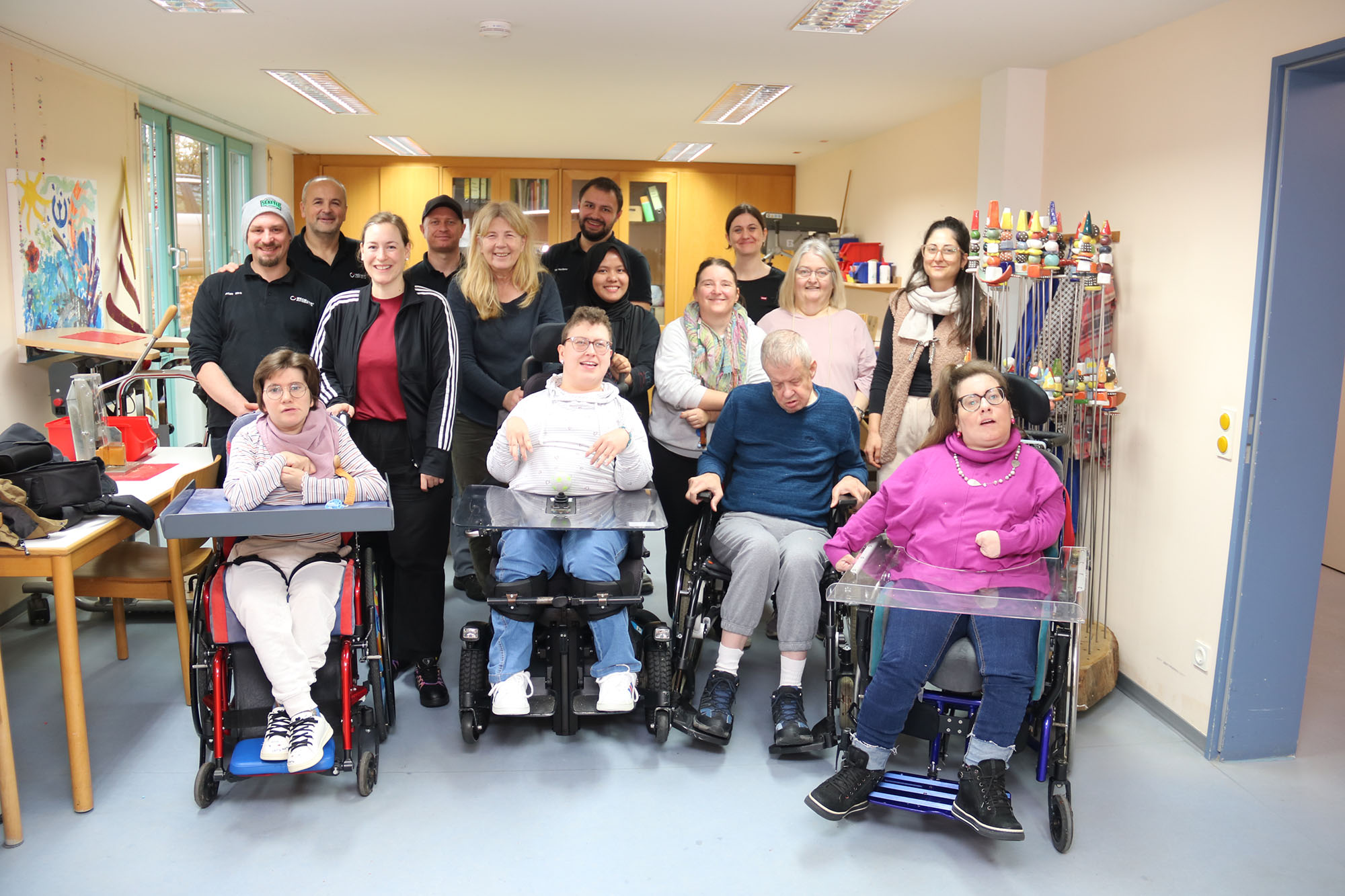 Eine Gruppe von Menschen steht im Halbkreis in einem Gruppenbild. Vorne sitzen drei Frauen und ein Mann in Rollstühlen.