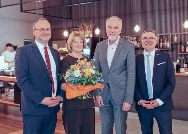 Vier Personen stehen nebeneinander in einem Raum. Eine Person hält einen großen Blumenstrauß mit orangefarbenen und weißen Blüten.