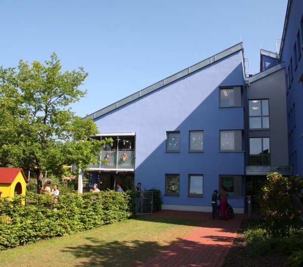 Ein blaues Haus in der Frontansicht. Vor dem Haus ist ein grüner Garten und ein großer Baum.