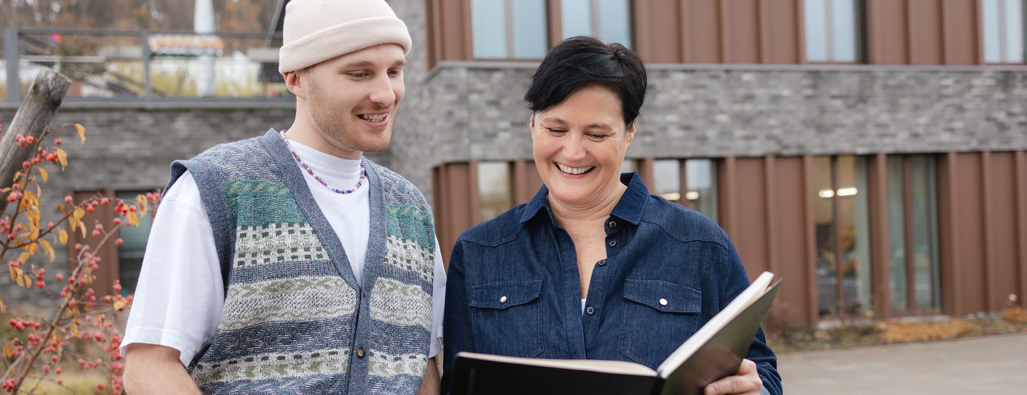 Ein Mann und eine Frau stehen draußen vor einem Gebäude. Die Frau hält ein offenes Buch in der Hand. Beide schauen in das Buch und lächeln.