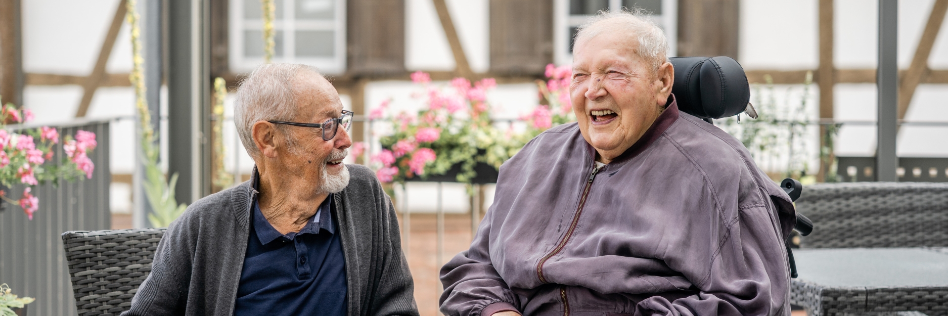 Zwei Senioren lachen gemeinsam. Einer der Senioren trägt eine Brille, der andere sitzt im Rollstuhl. Hinter ihnen ist ein Fachwerkhaus zu sehen. Sie befinden sich auf einem Balkon, der mit Blumen geschmückt ist. 