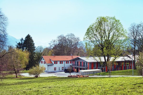 Auf einem Landschaftsfoto sieht man zwei Häuserseitlich. Im Vordergrund ist eine grüne Wiese, im Hintergrund Bäume, Berge und blauer Himmel zu sehen.