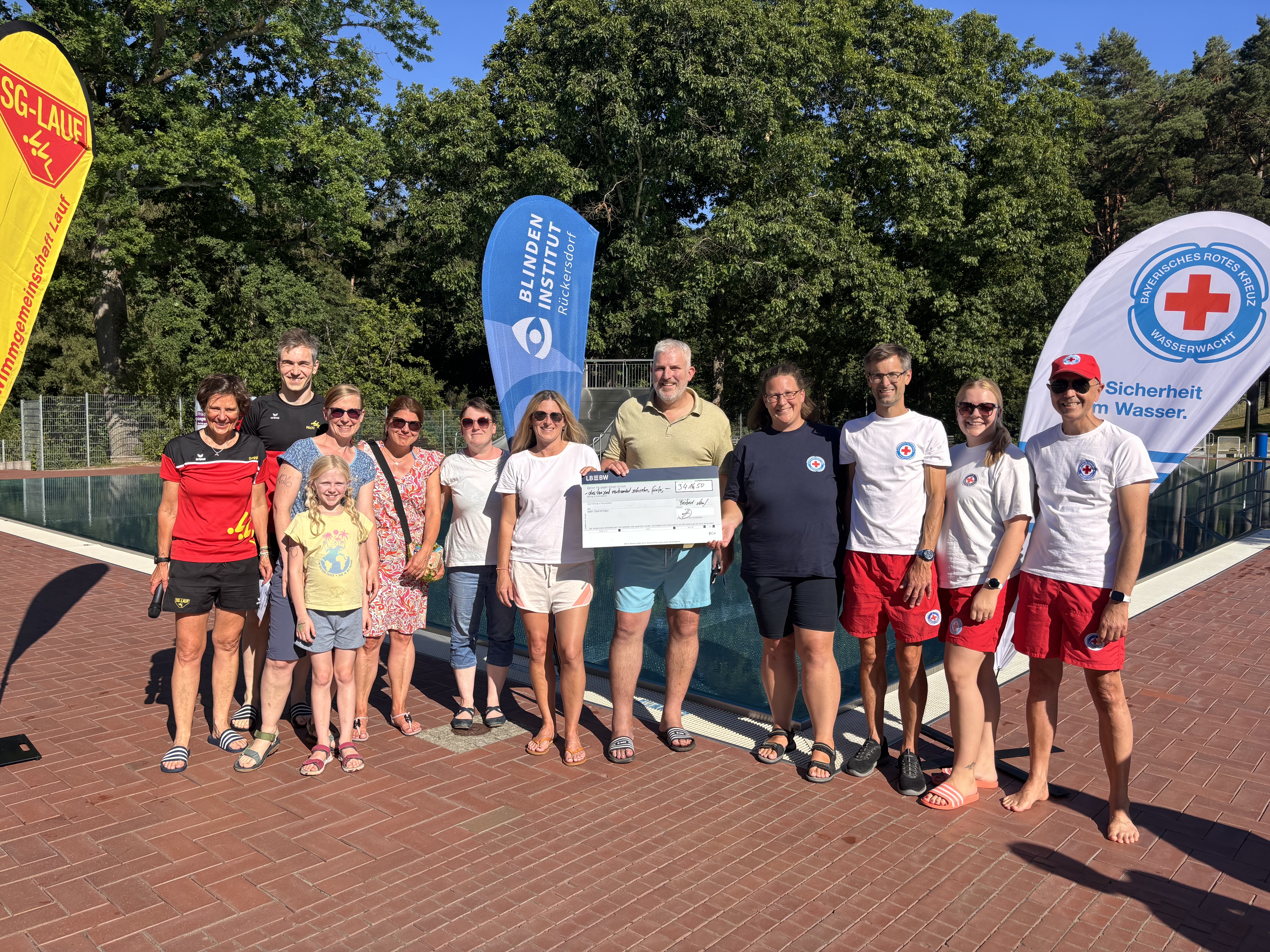 Scheckübergabe am Freibad Lauf: Vertreterinnen und Vertreter der Schwimmgemeinschaft Lauf, der Wasserwacht, des Blindeninstituts Rückersdorf sowie weitere Beteiligte stehen gemeinsam am Beckenrand. Im Hintergrund sind Fahnen des Blindeninstituts Rückersdorf, der SG Lauf und der Wasserwacht zu sehen.