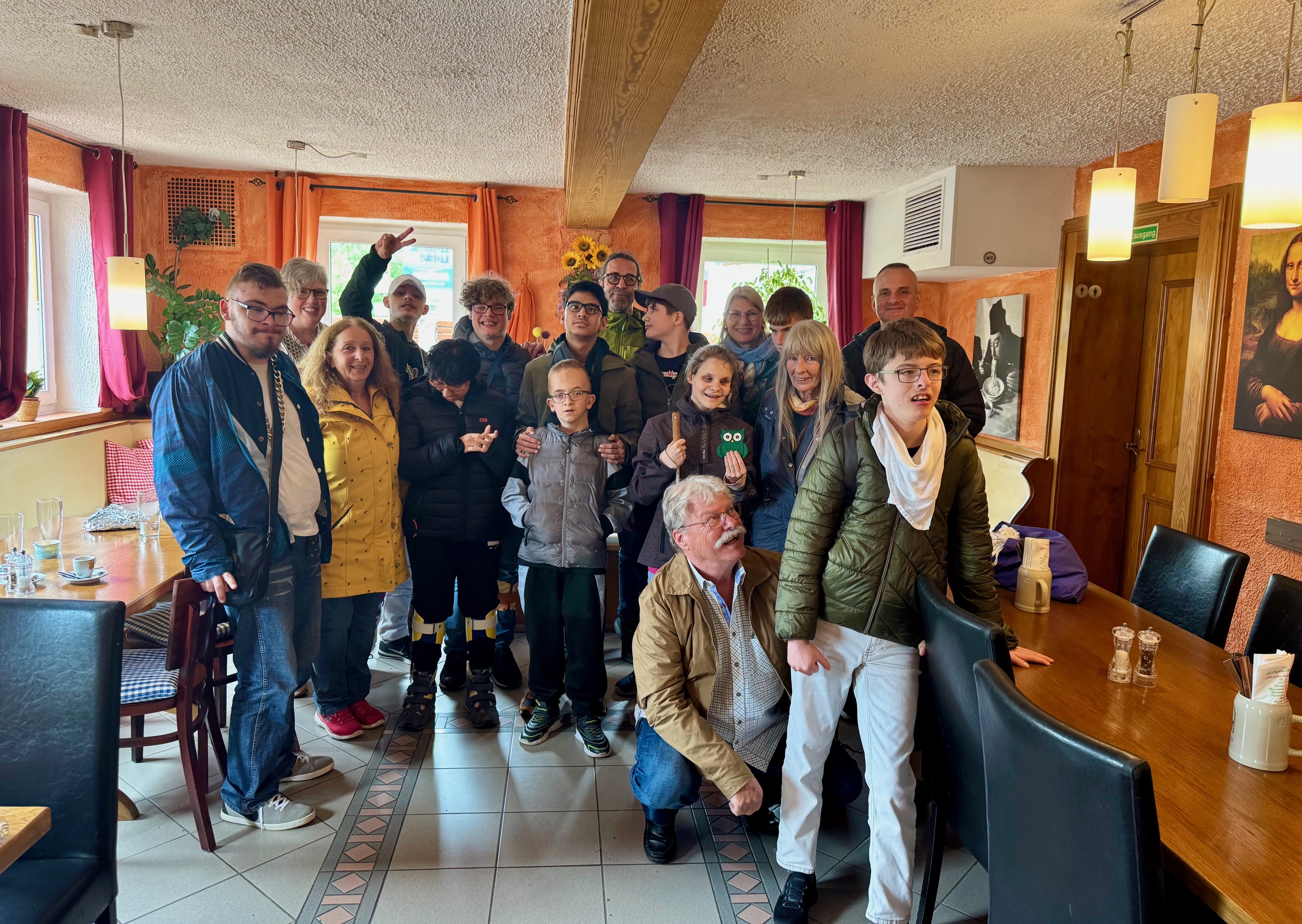 Gruppe von Jugendlichen und Erwachsenen aus der Schülerband Studio D in einer Pizzeria, umgeben von Holztischen und orangenen Wänden. Die Teilnehmenden stehen und sitzen dicht beieinander für ein Gruppenfoto, einige zeigen Handzeichen oder halten kleine Gegenstände; im Hintergrund sind Fenster, Pflanzen und Wandbilder zu sehen.