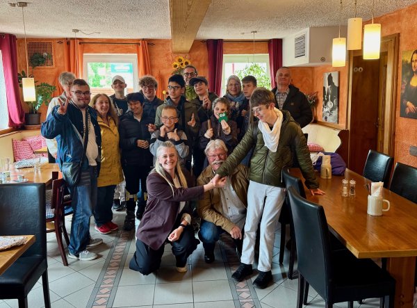 Gruppe von Jugendlichen und Erwachsenen aus der Schülerband Studio D in einer Pizzeria, umgeben von Holztischen und orangenen Wänden. Die Teilnehmenden stehen und sitzen dicht beieinander für ein Gruppenfoto, einige zeigen Handzeichen oder halten kleine Gegenstände; im Hintergrund sind Fenster, Pflanzen und Wandbilder zu sehen.