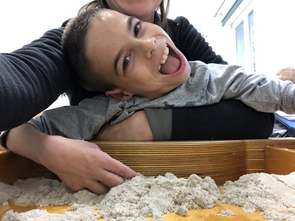 Auf dem Foto sind eine Frau und ein lachendes Kind zu sehen. Vor ihnen ist eine Holzkiste mit Knetmasse. Das Kind langt mit der Hand in die Knetmasse und die Frau umarmt dabei das Kind.