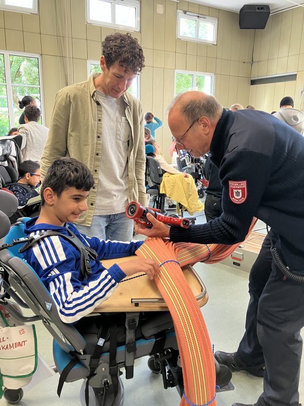 Das Bild zeigt einen Jungen beim Betasten eines Feuerwehrschlauchs. Neben ihm stehen zwei Männer und schauen dabei zu.