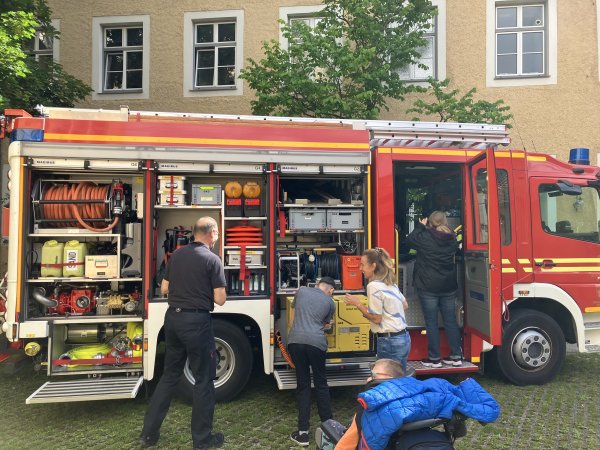 Das Bild zeigt einen Jungen im Rollstuhl, der einen Feuerwehrschlauch berührt. Neben ihm stehen zwei Männer.