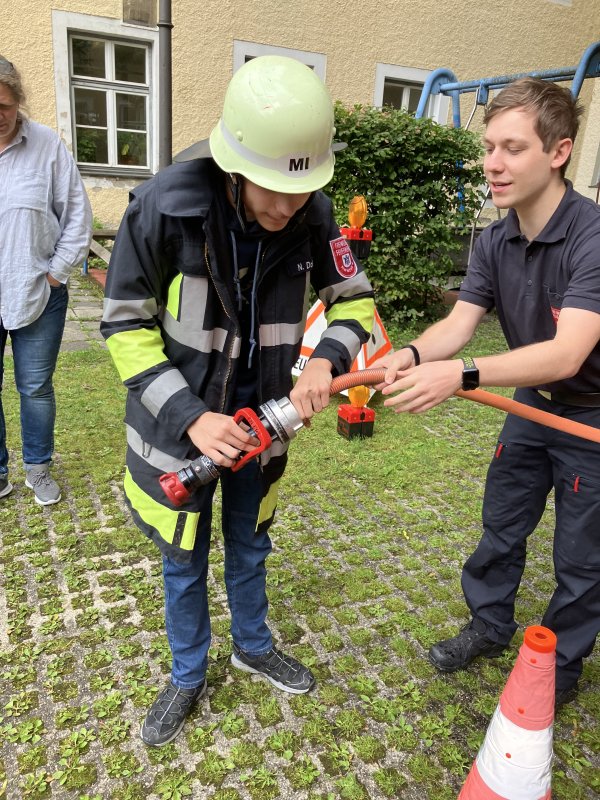 Das Bild zeigt einen Menschen in Feuerwehrmontur und mit einem Feuerwehrschlauch. Daneben steht ein junger Mann.