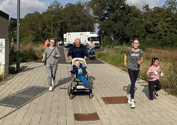 Zwei Erwachsene und drei Kinder laufen frontal auf den Betrachter zu. Ein Kind sitzt in einem Kinderwagen.