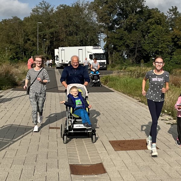 Zwei Erwachsene und drei Kinder laufen frontal auf den Betrachter zu. Ein Kind sitzt in einem Kinderwagen.