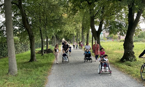 Eine Gruppe von Menschen, Erwachsene und Kinder, teils im Kinderwagen oder Rollstuhl, laufen auf einem Schotterweg, der von Bäumen eingegrenzt ist.