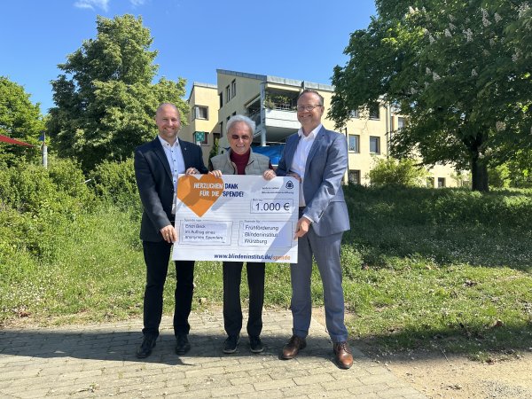 Drei Männer stehen auf dem Gelände des Blindeninstituts und haben einen Spendenscheck in der Hand.