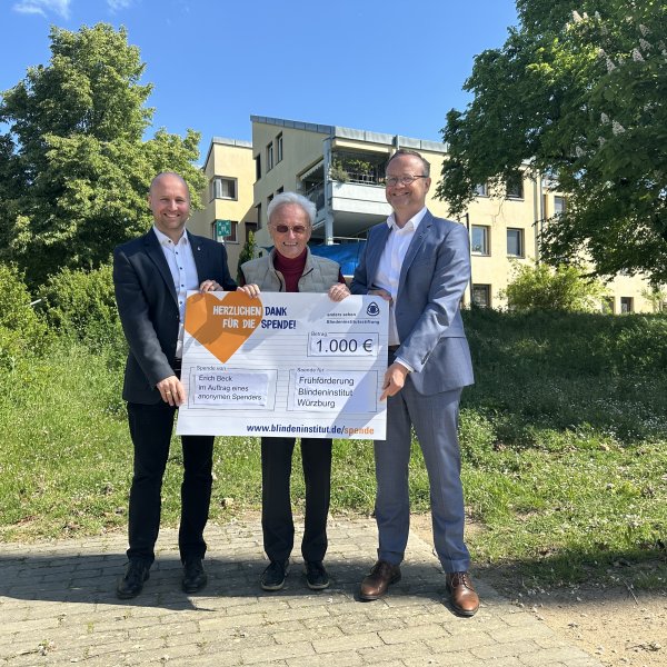 Drei Männer stehen auf dem Gelände des Blindeninstituts und haben einen Spendenscheck in der Hand.