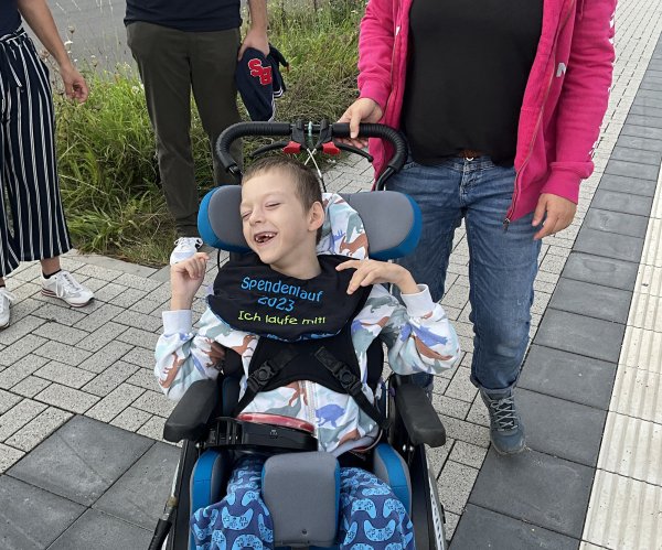 Ein kleiner Junge im Rollstuhl lacht in die Kamera, er trägt ein T-Shirt mit Aufdurck Spendenlauf 2023 - ich laufe mit.