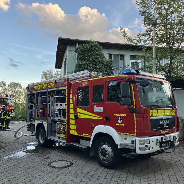 Ein Einsatzfahrzeug der Freiwilligen Feuerwehr Rückersdorf steht vor einem Gebäude des Blindeninstituts Rückersdorf. Mehrere Feuerwehrkräfte sind vor Ort, Schläuche liegen auf dem Boden. Im Hintergrund ist ein weiteres Feuerwehrfahrzeug zu sehen. Die Aufnahme entstand während einer Rettungsübung.