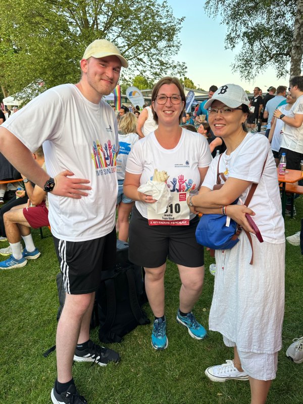 Ein Mann und zwei Frauen im Sport-Outfit stehen auf einer Wiese und lachen. Im Hintergrund stehen weitere Sportler*innen oder sitzen auf Bierbänken.