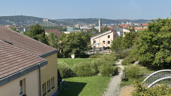 Dach eines Gebäudes im Vordergrund, dahinter eine Grünfläche mit Spielplatz und einer Stadtansicht mit Gebäuden, einem Schloss und bewaldeten Hügeln unter blauem Himmel.