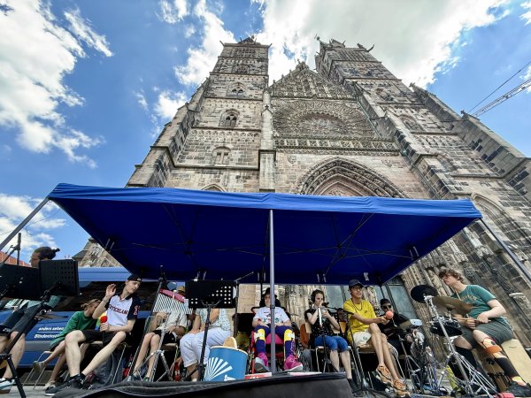 Eine Gruppe von Jugendlichen und Erwachsenen aus der Schülerband Studio D sitzt unter einem blauen Pavillon vor der beeindruckenden Fassade der Lorenzkirche Nürnberg, einer großen gotischen Kirche mit zwei Türmen. Die Personen spielen verschiedene Instrumente wie Trommel, Schlagzeug und Klangstäbe. Im Vordergrund sind Notenständer und Mikrofone zu sehen, im Hintergrund ragt die detailreiche Steinfassade der Kirche vor blauem Himmel mit Wolken auf.